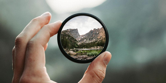 Eine Hand hält eine Linse aus Glas vor unscharfem Hintergrund, durch die eine Berglandschaft zu sehen ist